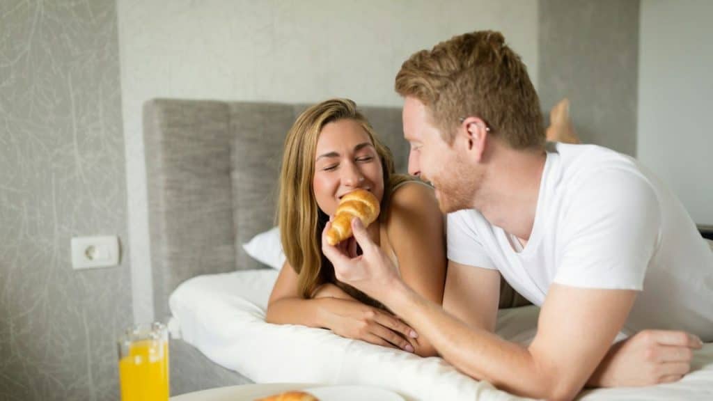 Man feeds a croissant to a woman while they relax together in bed.