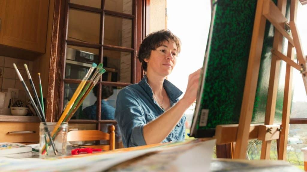 Middle-aged woman with short hair smiles while painting on an easel indoors near a window.