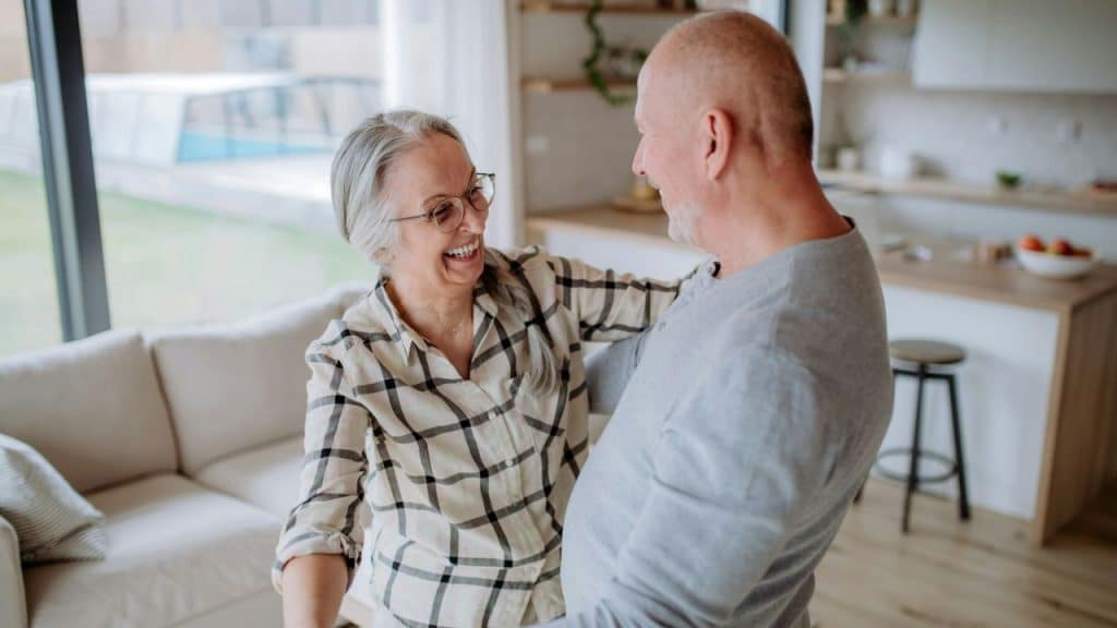 Smiling elderly couple dances and embraces in a bright, modern living room.