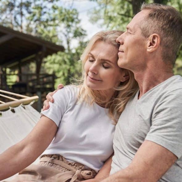 Middle-aged couple relaxing in a hammock outside a wooden house on a sunny day.