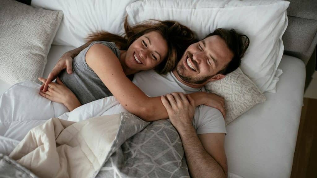 Smiling couple embracing each other while lying down together in a white bed.