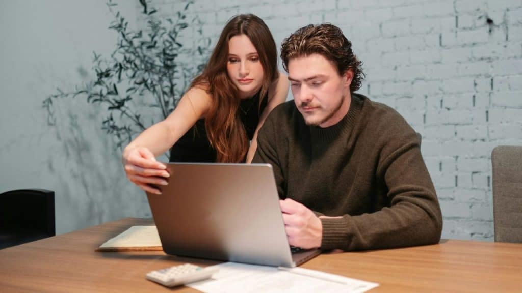 A man and woman are focused on a laptop screen together at a wooden table.