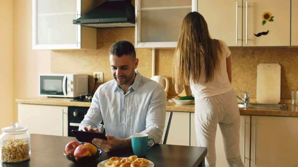 A man sits at a table using a tablet while a woman works in the kitchen behind him.