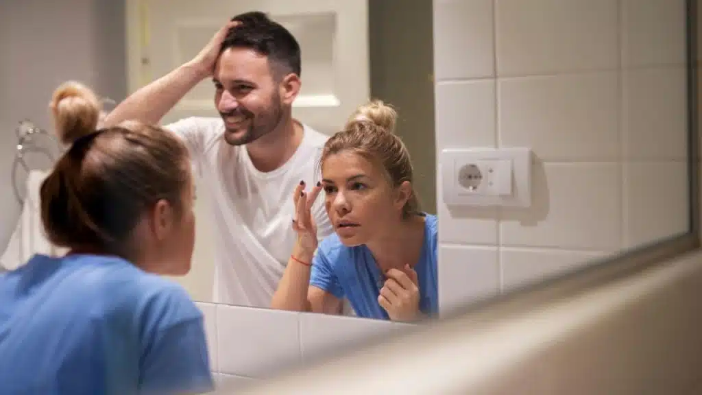 A man and woman look at their reflections in a bathroom mirror together.