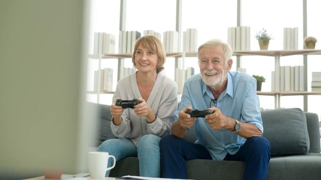 Smiling older couple sitting on a couch, holding controllers and playing video games.