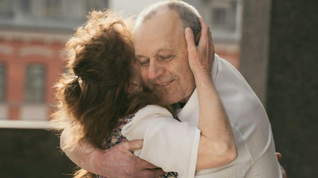 Older couple embracing warmly outdoors with the man smiling and eyes closed.