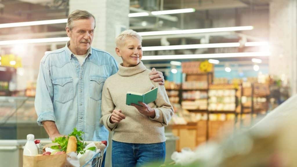 Older couple shopping in a brightly lit grocery store, checking a small notebook.