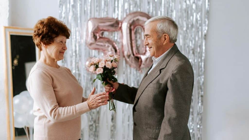 Older man giving a bouquet of roses to an older woman in front of a "50" balloon sign.