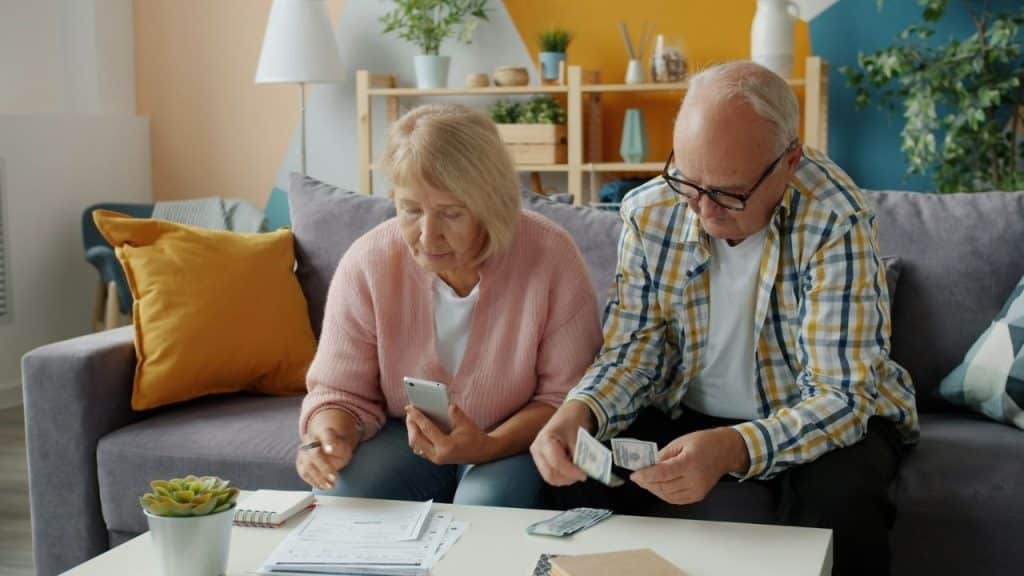 Older couple on a couch looking at a phone and counting cash and documents.