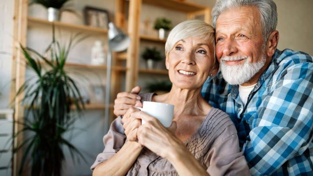 Older couple embracing indoors, smiling while the woman holds a white cup.