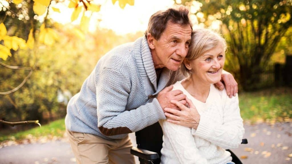 Older man embracing a smiling older woman sitting in a wheelchair outdoors in autumn.