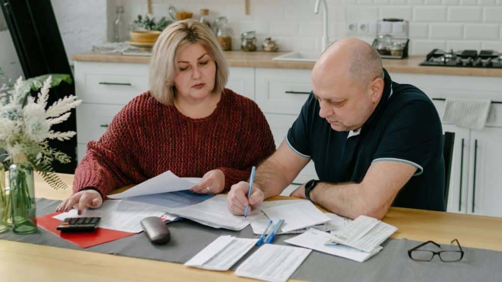 A middle-aged couple is sitting at a kitchen table, intently reviewing and signing papers.