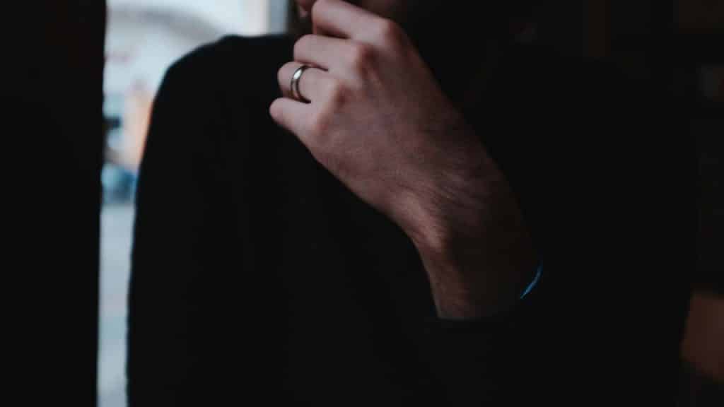 A close-up of a man’s hand with a ring resting near his face in a dim setting.