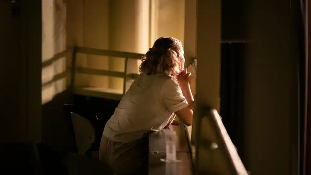 A woman sitting alone in dim light, gazing thoughtfully out a window.
