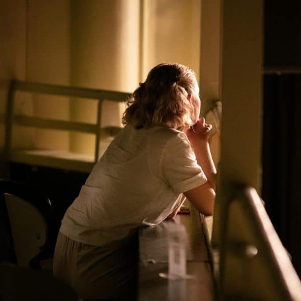 A woman sitting alone in dim light, gazing thoughtfully out a window.