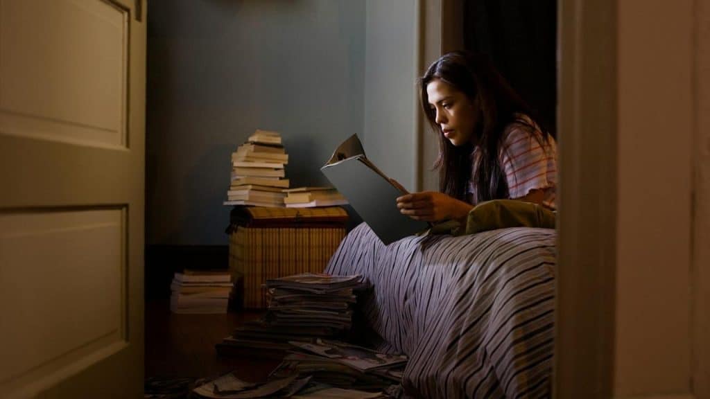 A woman sitting on a bed reading a book in a dim room surrounded by stacks of books.