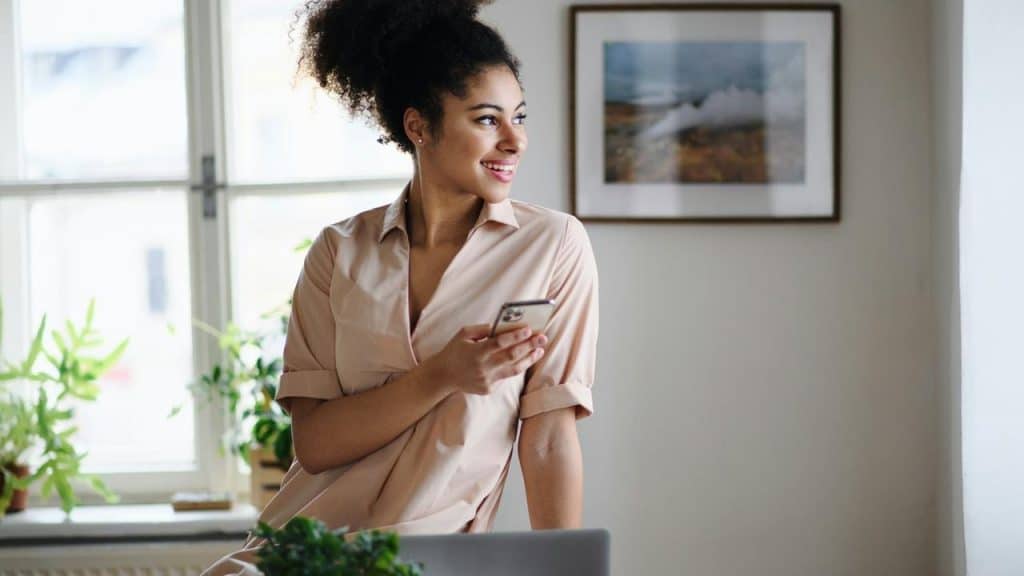 A smiling woman holding a smartphone looks out of a window while sitting on a desk.