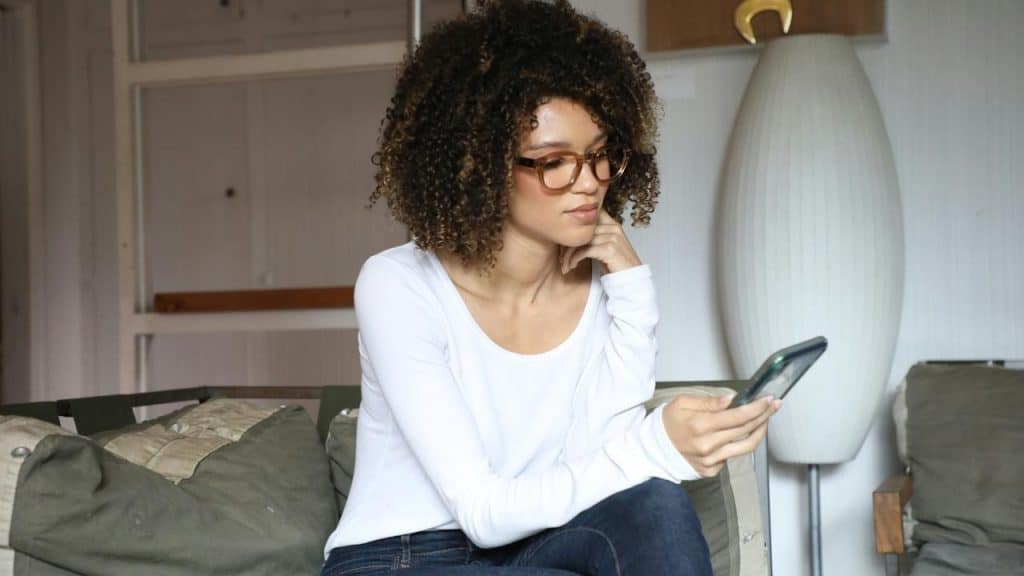 A woman wearing glasses sits on a couch, looking at her phone.
