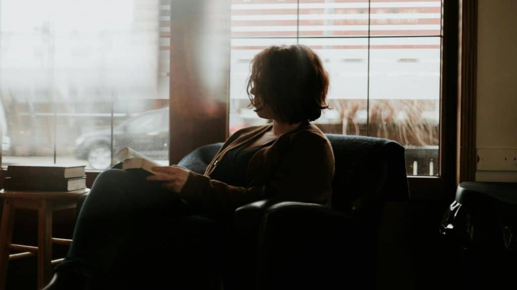 A woman sitting in a chair reading a book and gazing out a window.