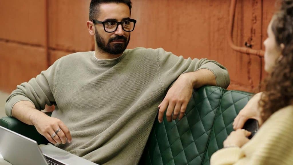 A man wearing glasses sits on a green couch talking to a woman.