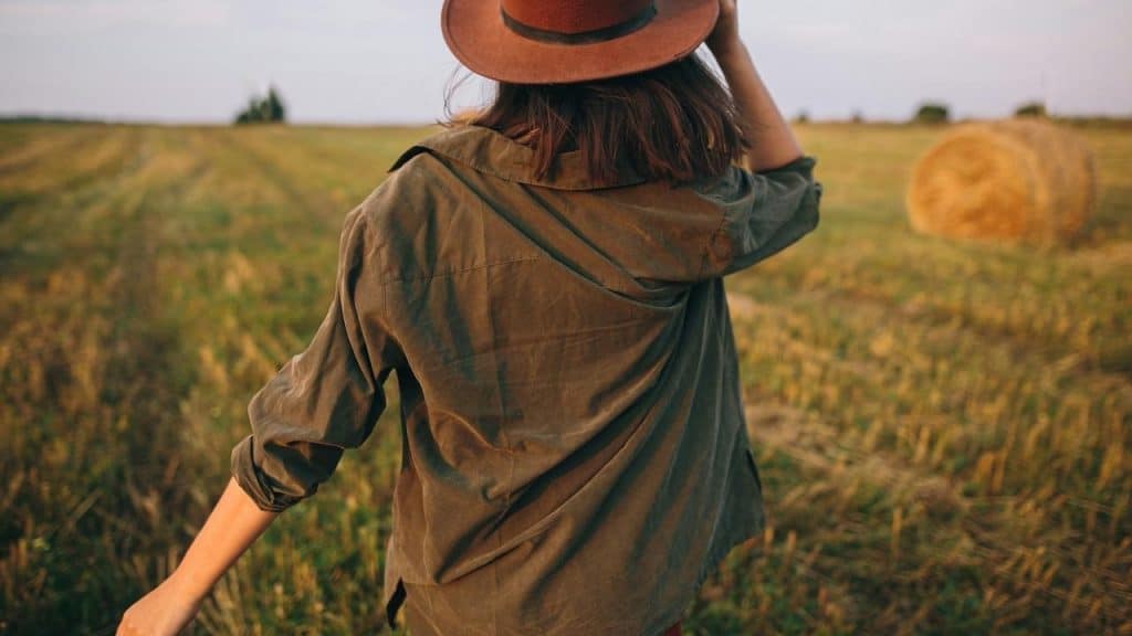 A person wearing a hat and green jacket walks through a field near a hay bale.