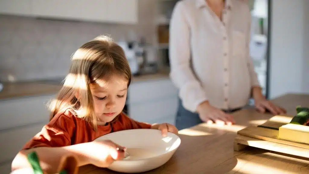 A little girl eating from a bowl at the kitchen table with her mother nearby.