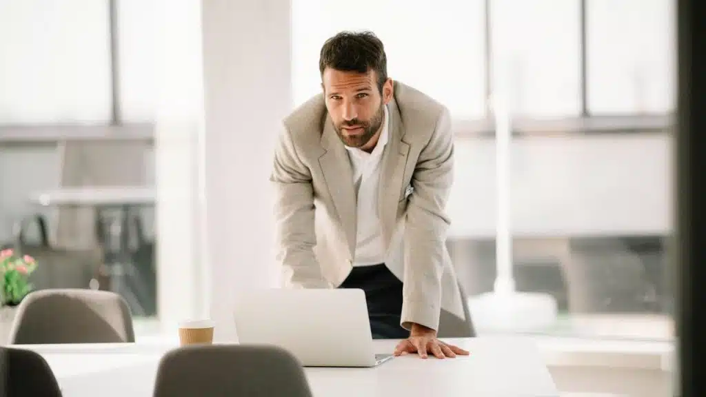 A man in a beige blazer leaning on a table with a laptop in front of him.