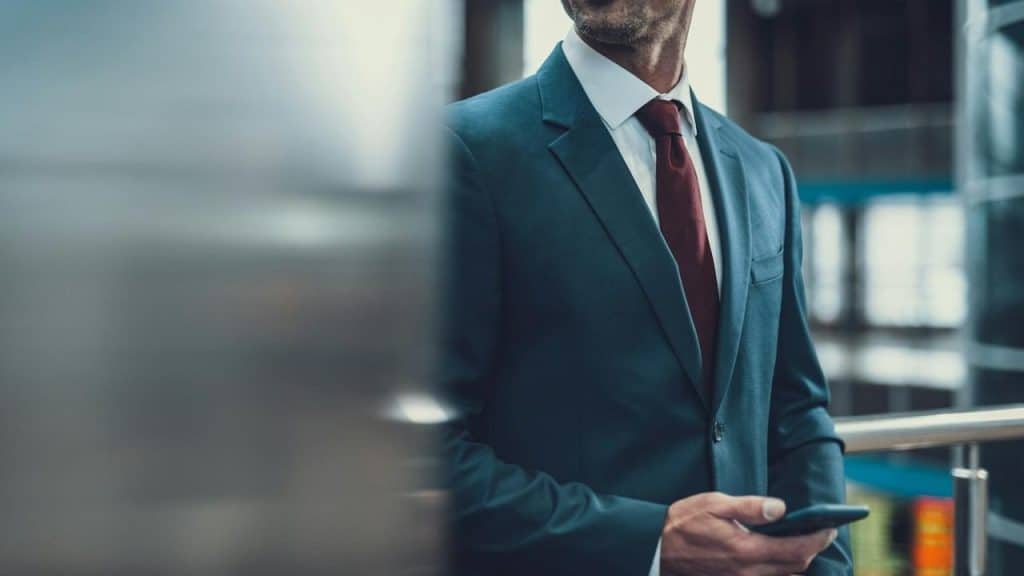 A man in a dark suit with a red tie holding a smartphone indoors.