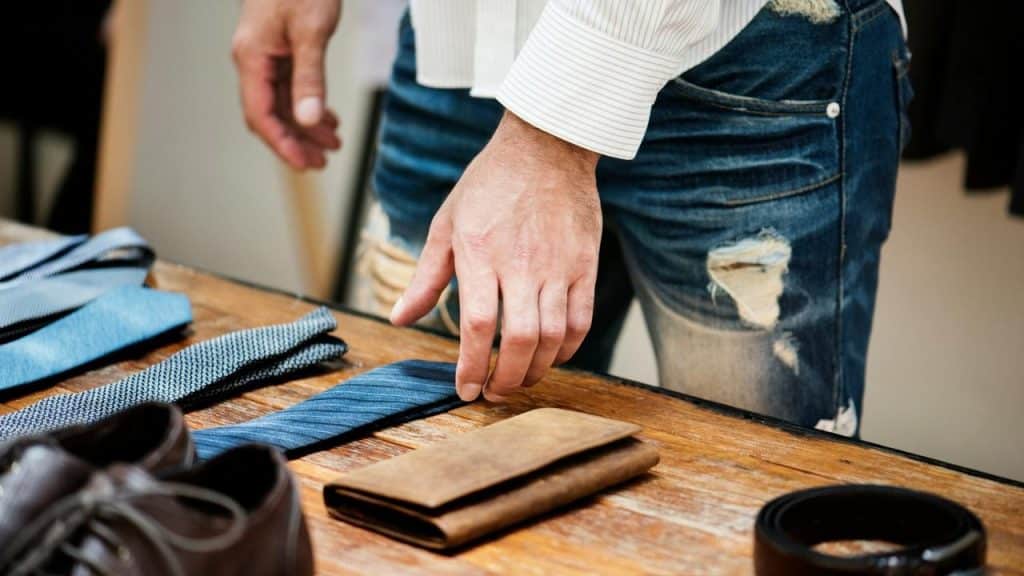 A person selecting ties on a wooden table next to shoes and a wallet.