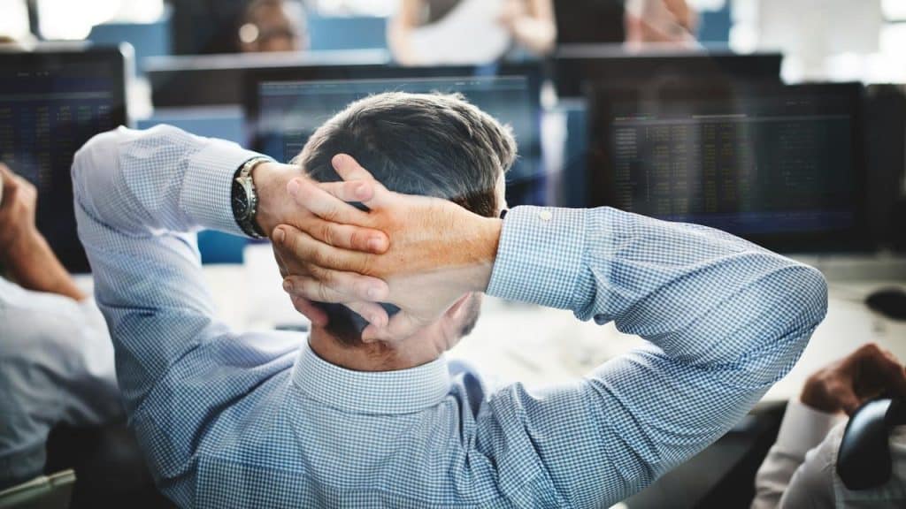 A person leaning back with hands behind their head at computer screens.