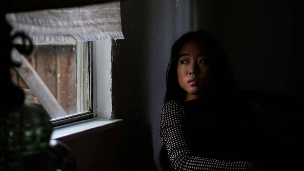 A woman sitting in a dim room looking toward a window.