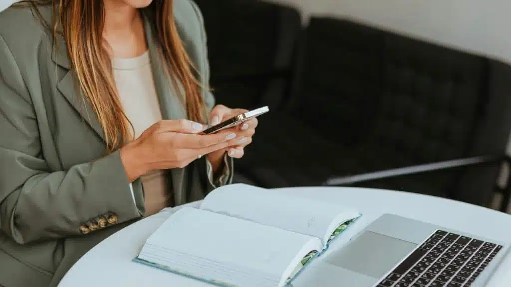 A woman using her phone beside an open notebook and laptop.