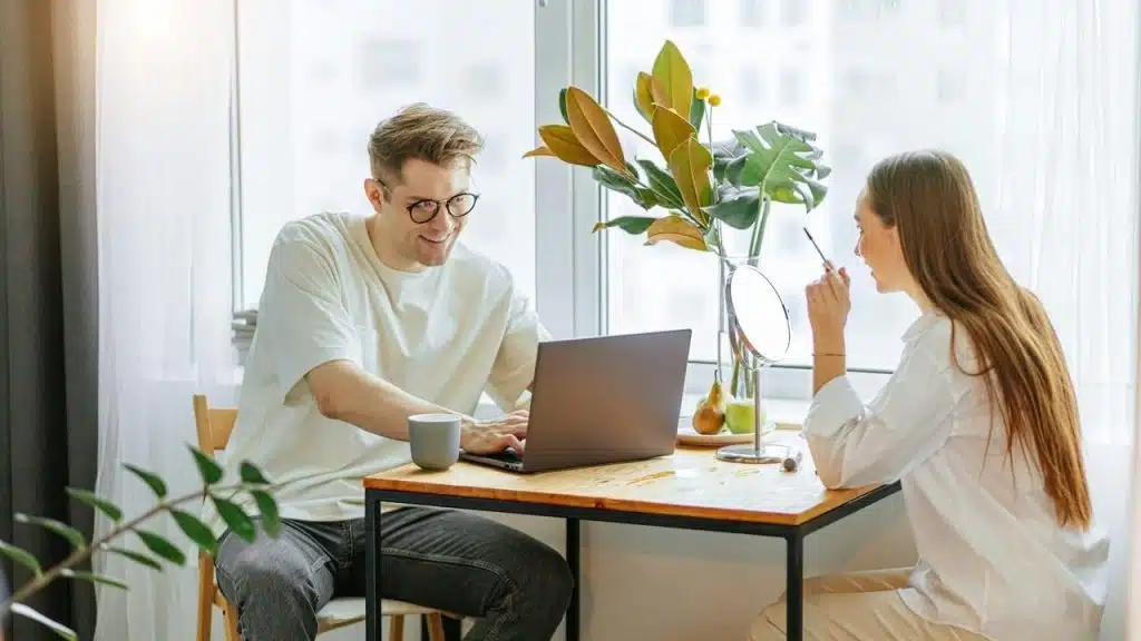 A man working on a laptop at a table while a woman sits across from him.