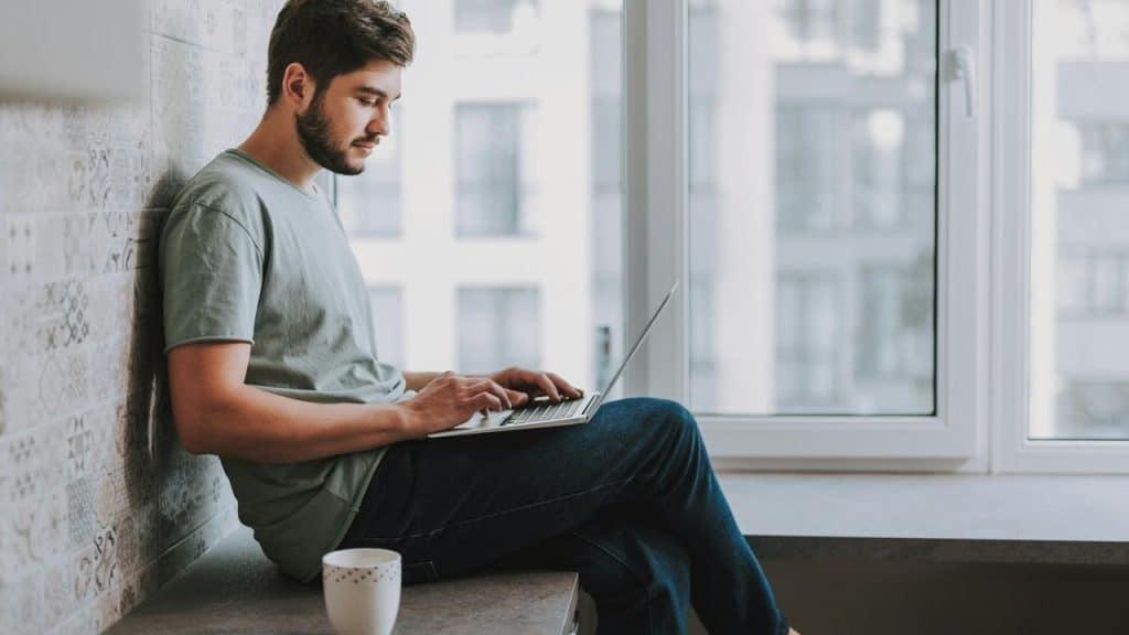 A man sitting by a window working on a laptop with a coffee mug nearby.