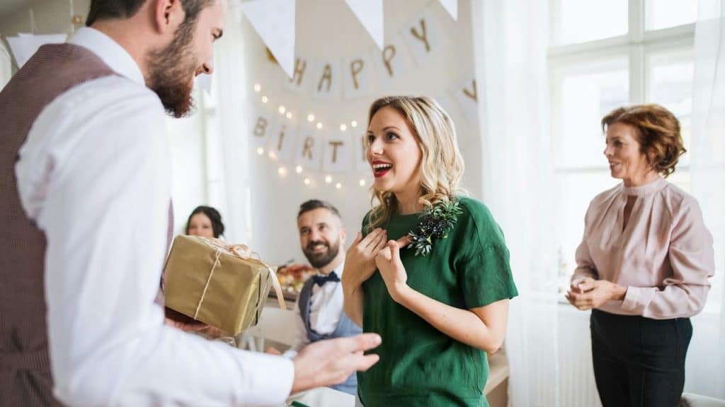 A woman smiling with surprise as she receives a wrapped birthday gift at a party.