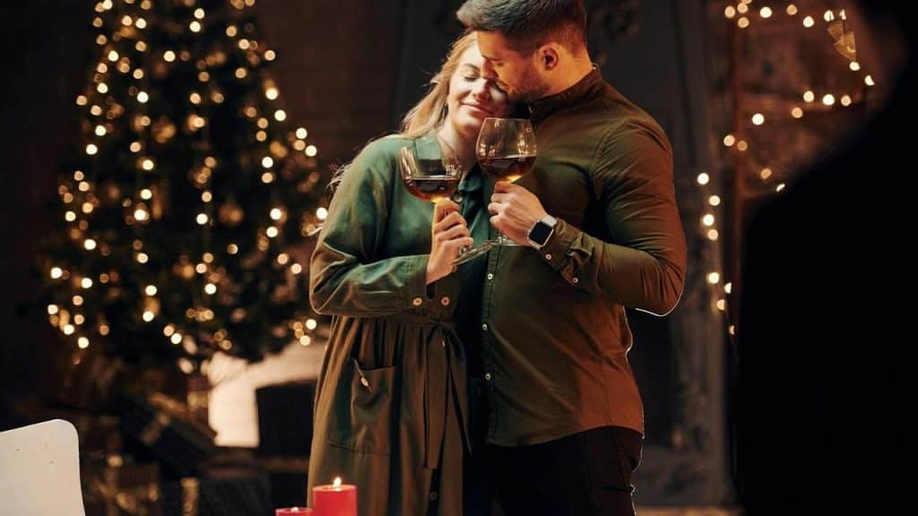 A couple holding wine glasses and embracing in front of a Christmas tree with lights.