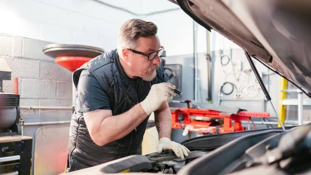 A mechanic inspecting a car engine with a flashlight in a garage.