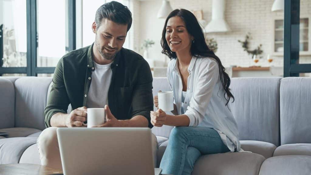 A couple sitting on a couch with coffee mugs, smiling while looking at a laptop.