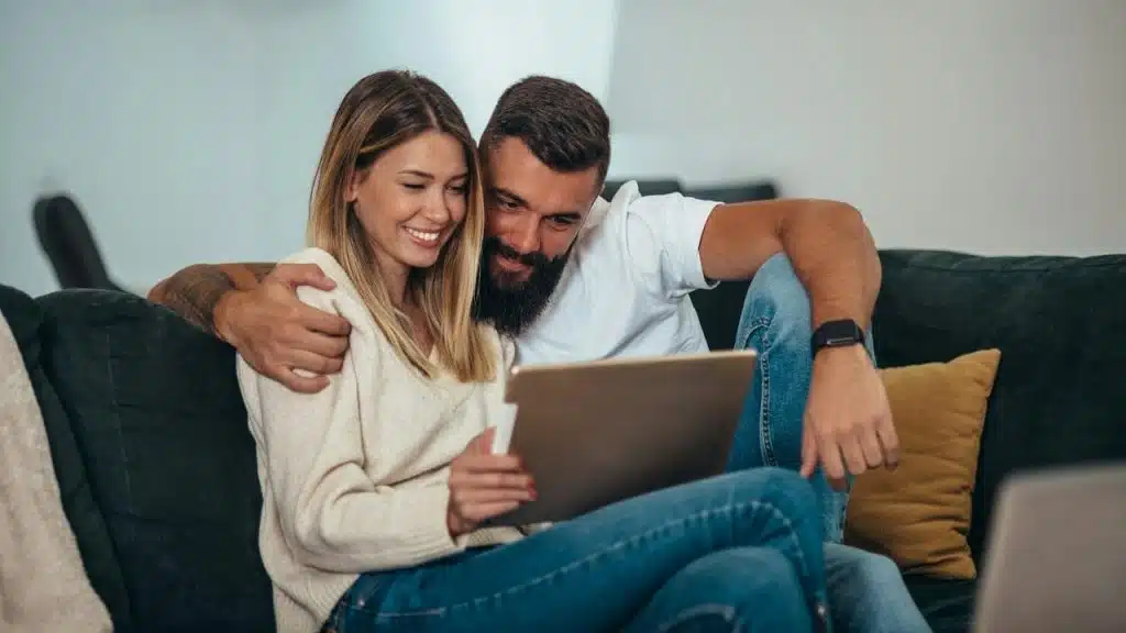 A couple sitting on a couch smiling while looking at a tablet.