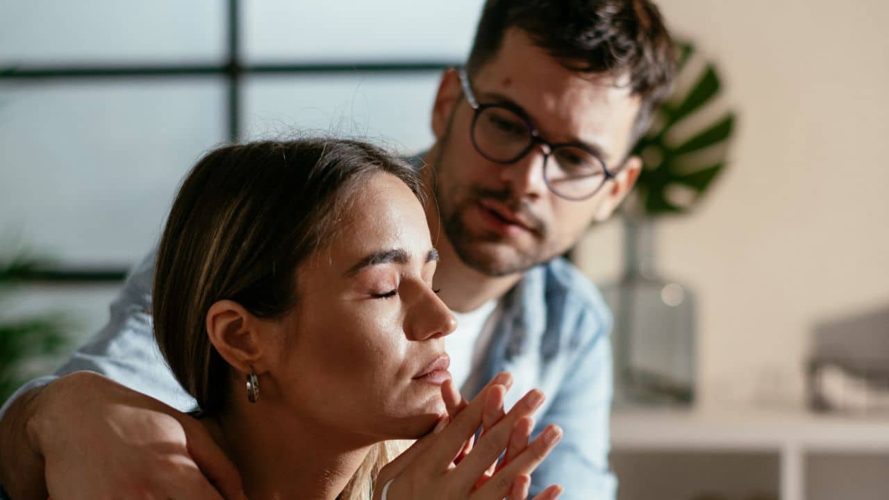 A man with glasses holds a woman's shoulders as she sits with her eyes closed.