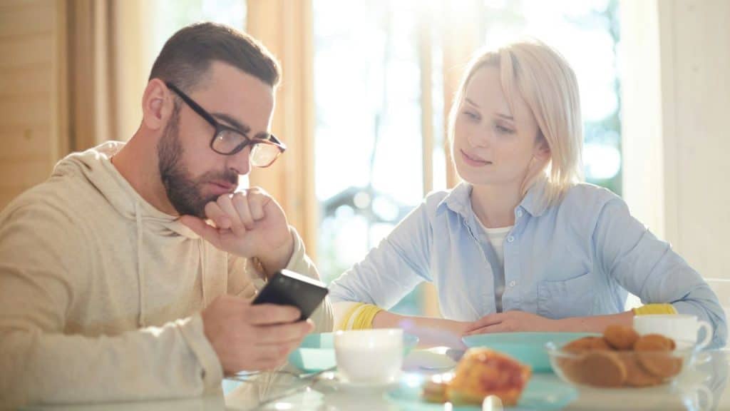 A man is absorbed in his phone while a woman smiles slightly, looking at him.