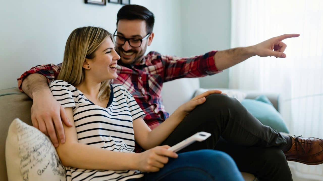 Smiling couple relaxing on a couch, with the man pointing and the woman holding a remote.