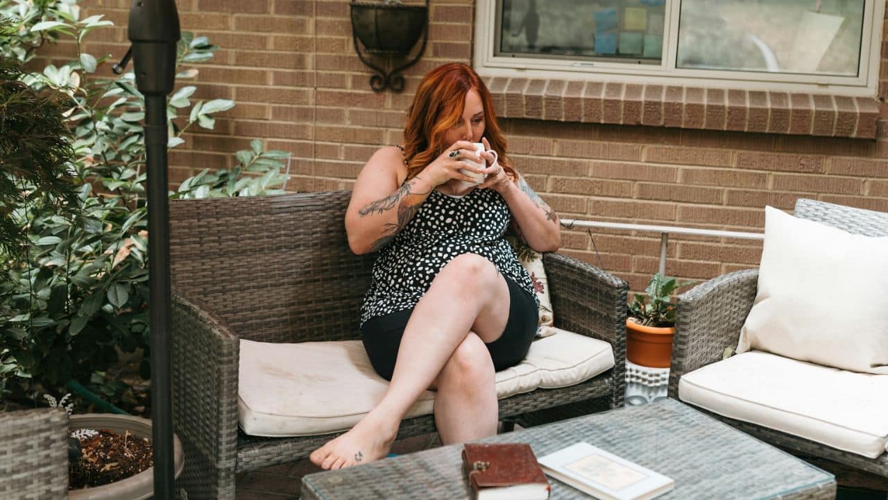 Woman with red hair sitting on a patio sofa, drinking from a white mug.