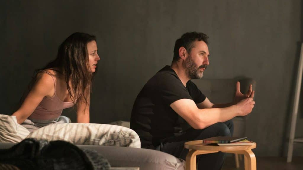 A man sits on the floor talking with hand gestures while a woman sits in bed.