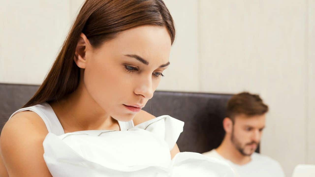 Close-up of a woman in a white tank top holding a pillow, with a blurry man in the background.