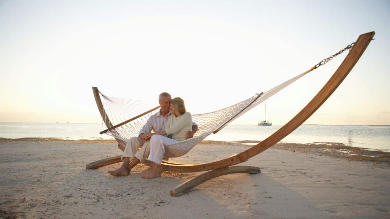 Older couple sitting on a wooden-framed hammock on a sunny beach near the ocean.