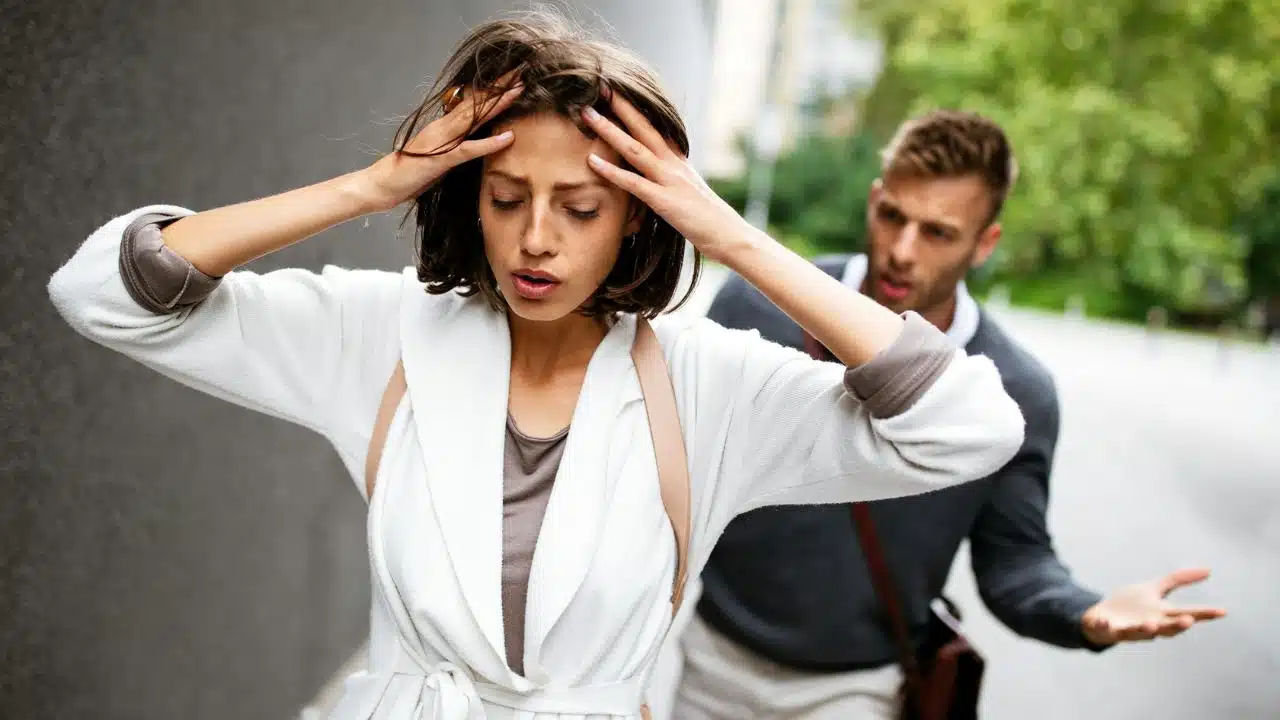 Woman holding her head in distress while a man gestures dismissively behind her.