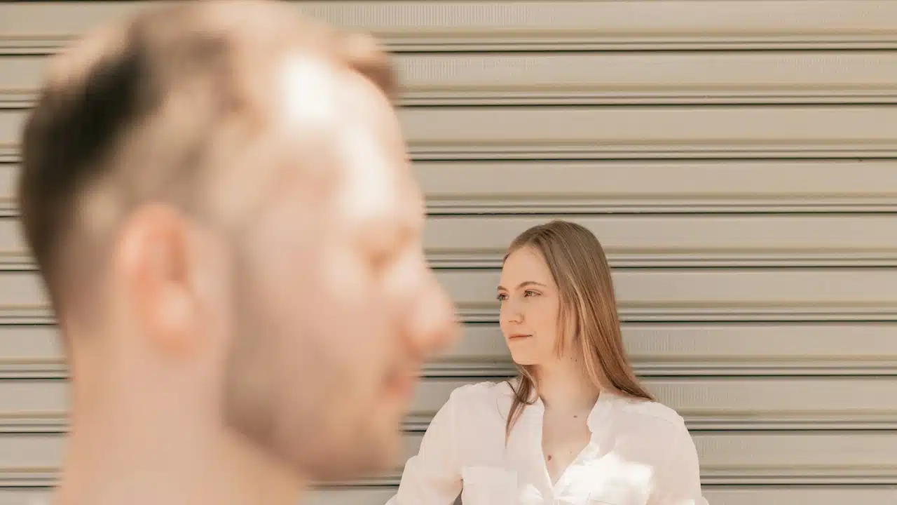 A woman looking to the side while leaning against a wall, with a man in the foreground. 