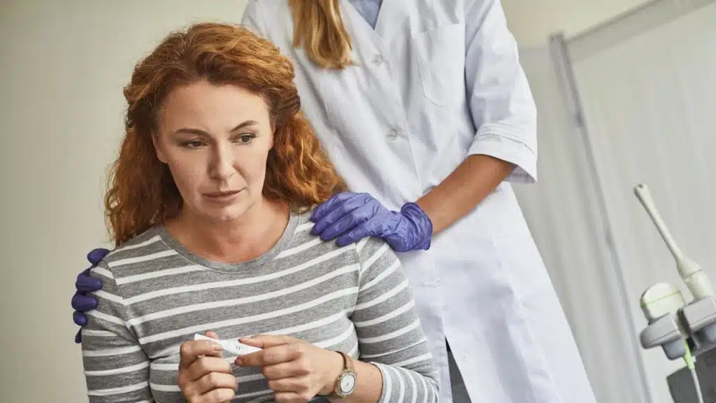An unhappy red-haired woman holding a pregnancy test with a negative result, with a supportive doctor comforting the lady and touching her shoulders.