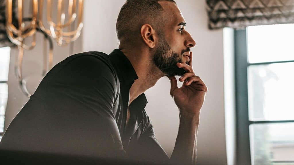 A man sits indoors with his hand on his chin, looking thoughtful.